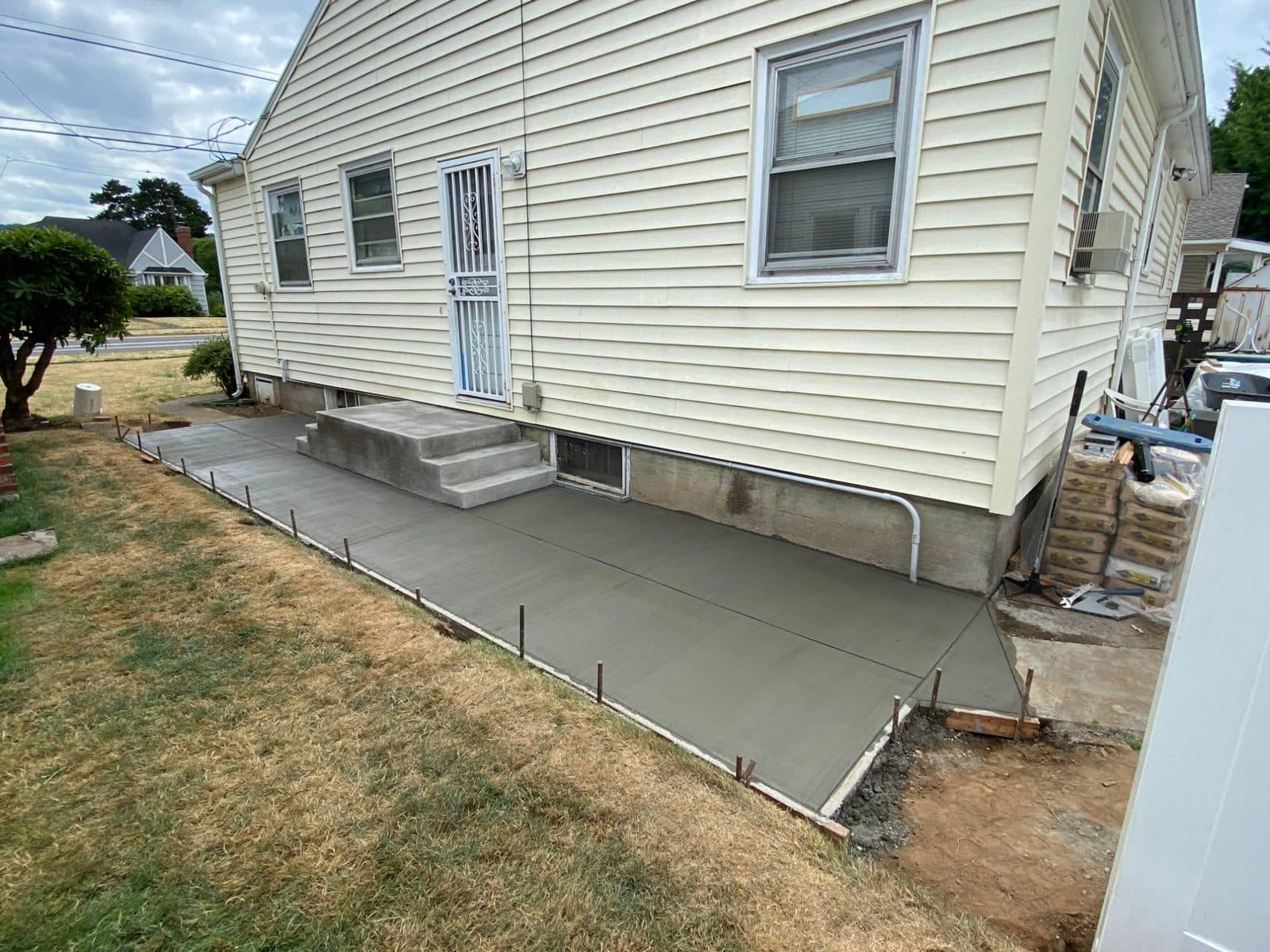 Acera de concreto recién vaciada y escalones de entrada de concreto nuevos a lo largo del costado de una casa con revestimiento color crema, con los moldes todavía puestos
