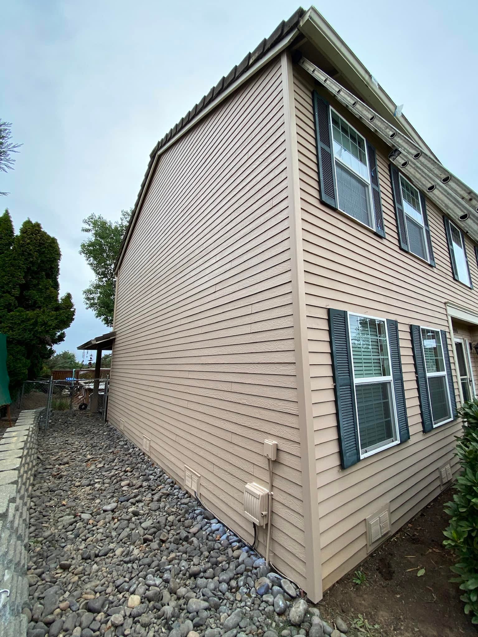 Tan-sided two-story house corner with dark shutters, river-rock bed along the foundation
