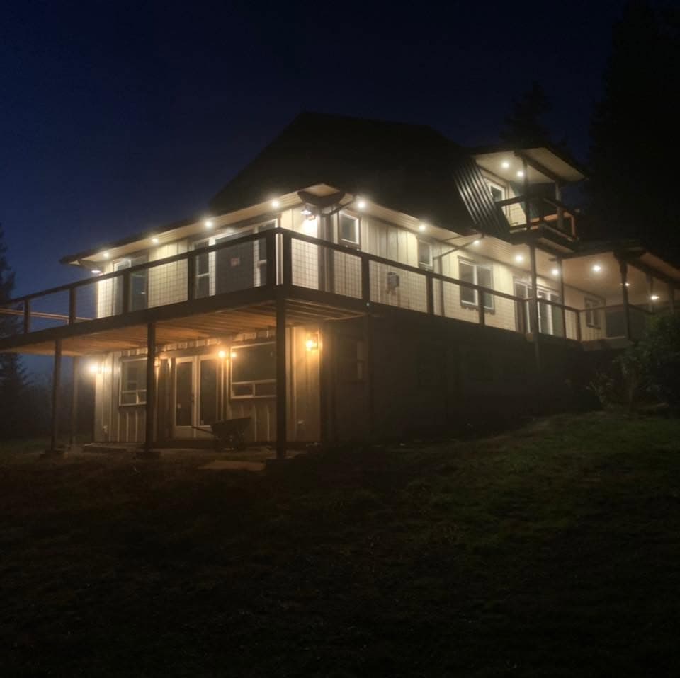 Casa de dos pisos de noche con una terraza elevada grande, barandal y un patio cubierto abajo, con las luces exteriores encendidas