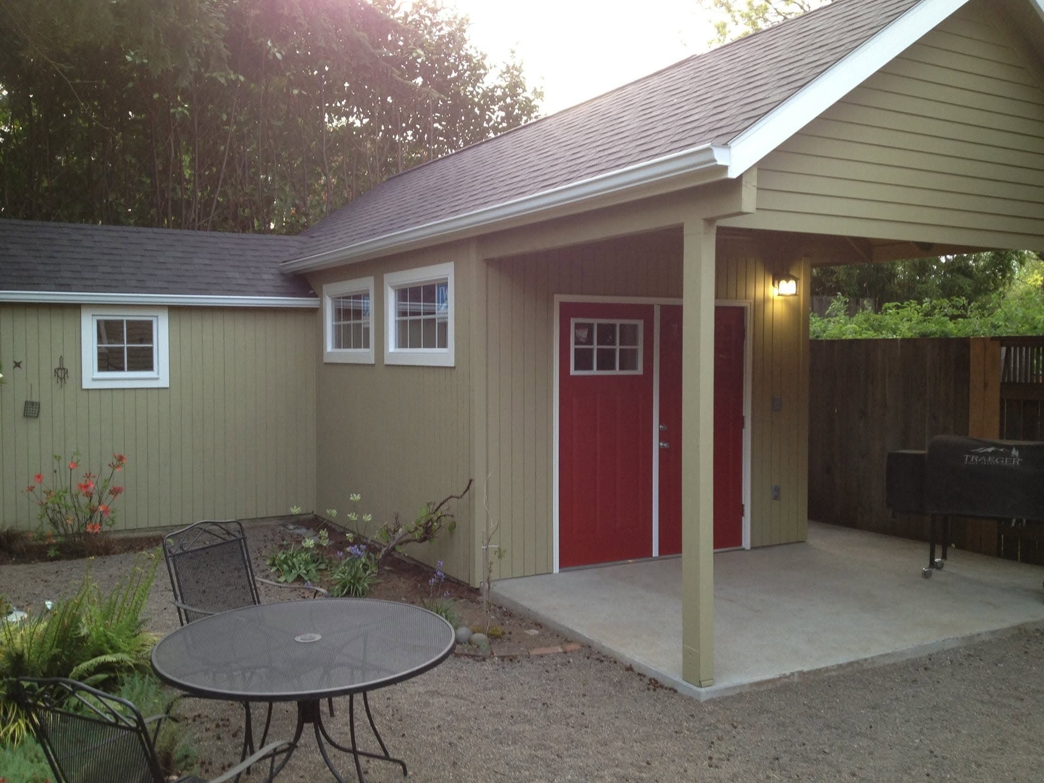 Finished backyard outbuilding with a covered patio, tan siding, and red double doors, patio table and chairs in front