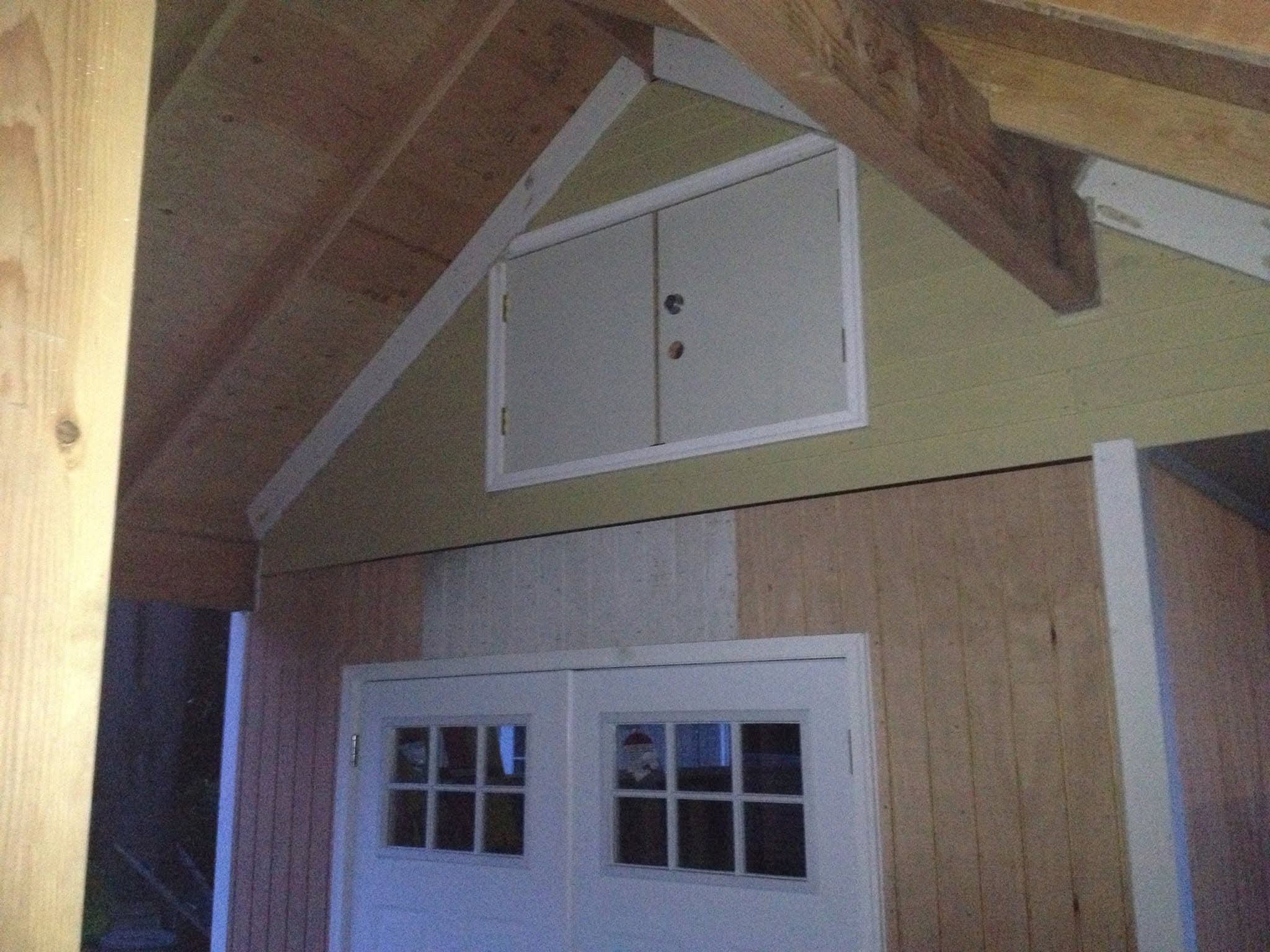 Interior of a wood-framed shop showing exposed rafters, small white cupola vent doors, and a paneled garage door below