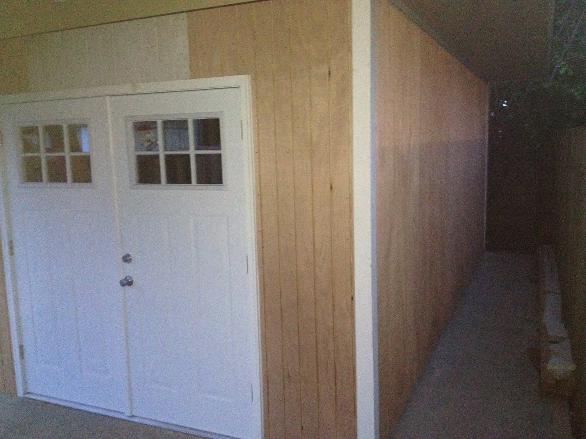 Interior de un taller con armazón de madera, con una puerta de garaje blanca con paneles y ventanas, y paredes de madera contrachapada (plywood) sin terminar