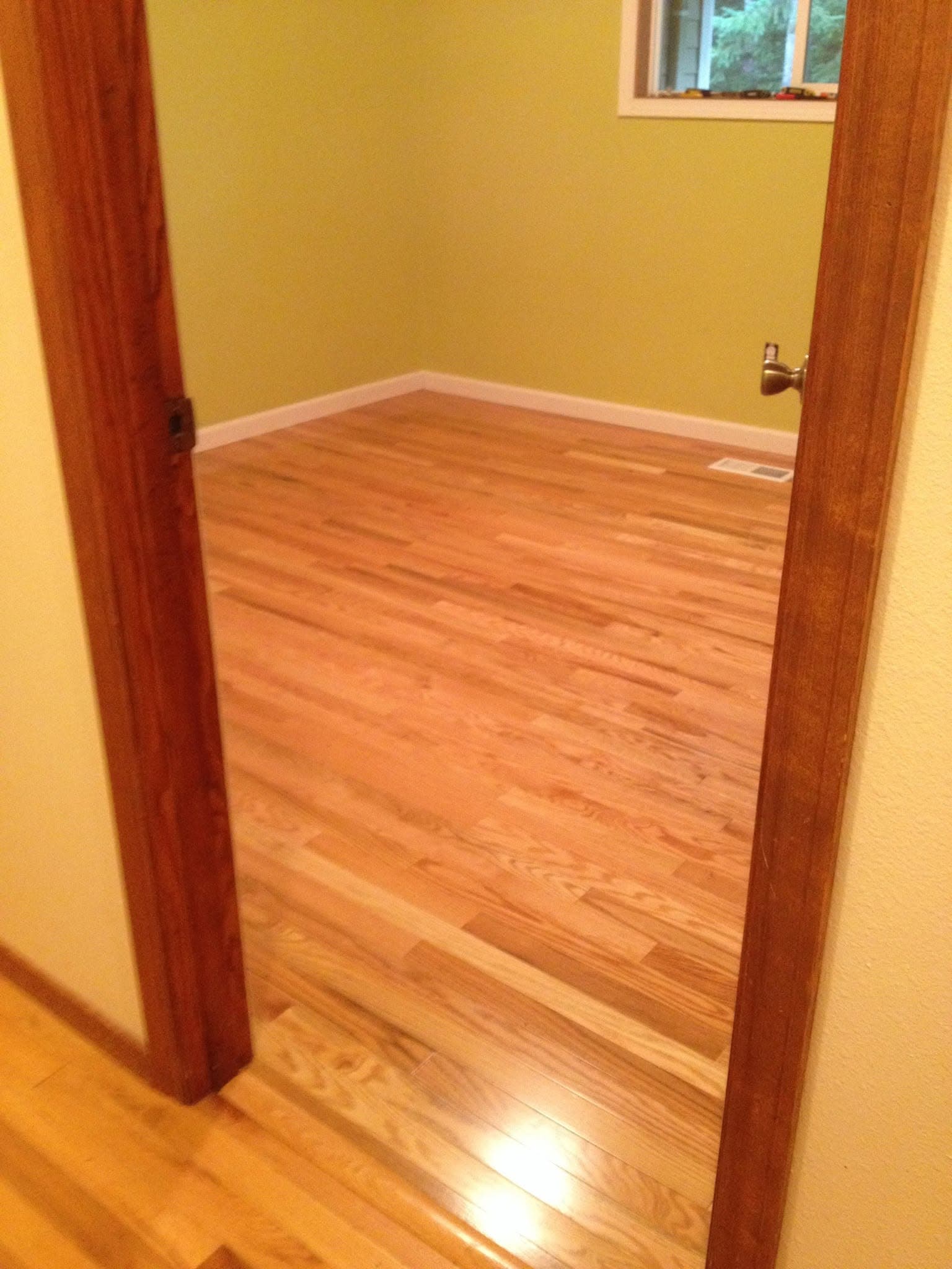 Piso de madera restaurado en un cuarto con paredes verde olivo y una ventana, con la puerta en primer plano