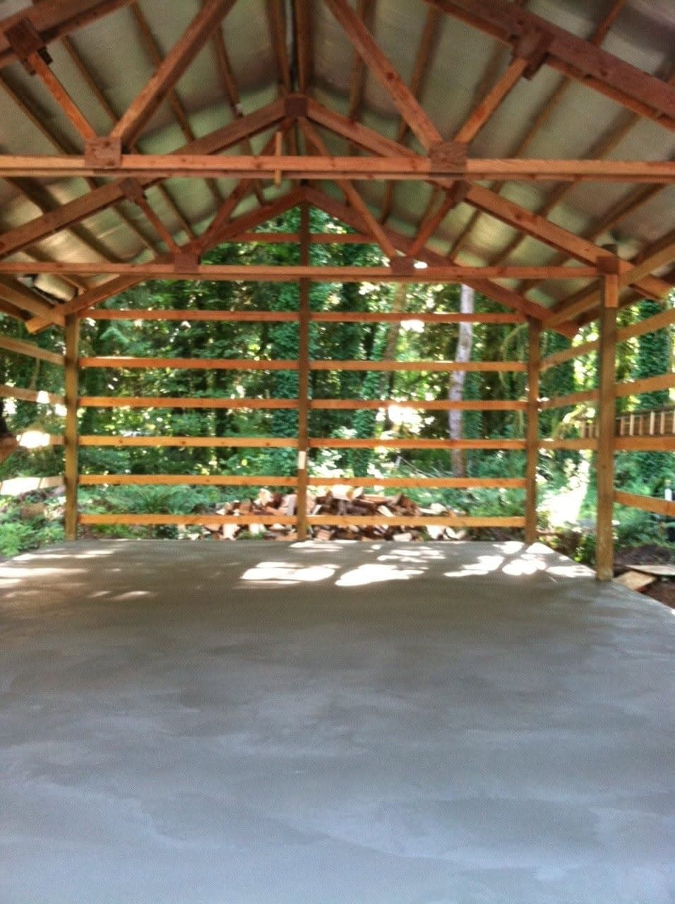 Pole barn abierto con armazón de madera, losa de piso de concreto terminada y techo de metal, con árboles atrás