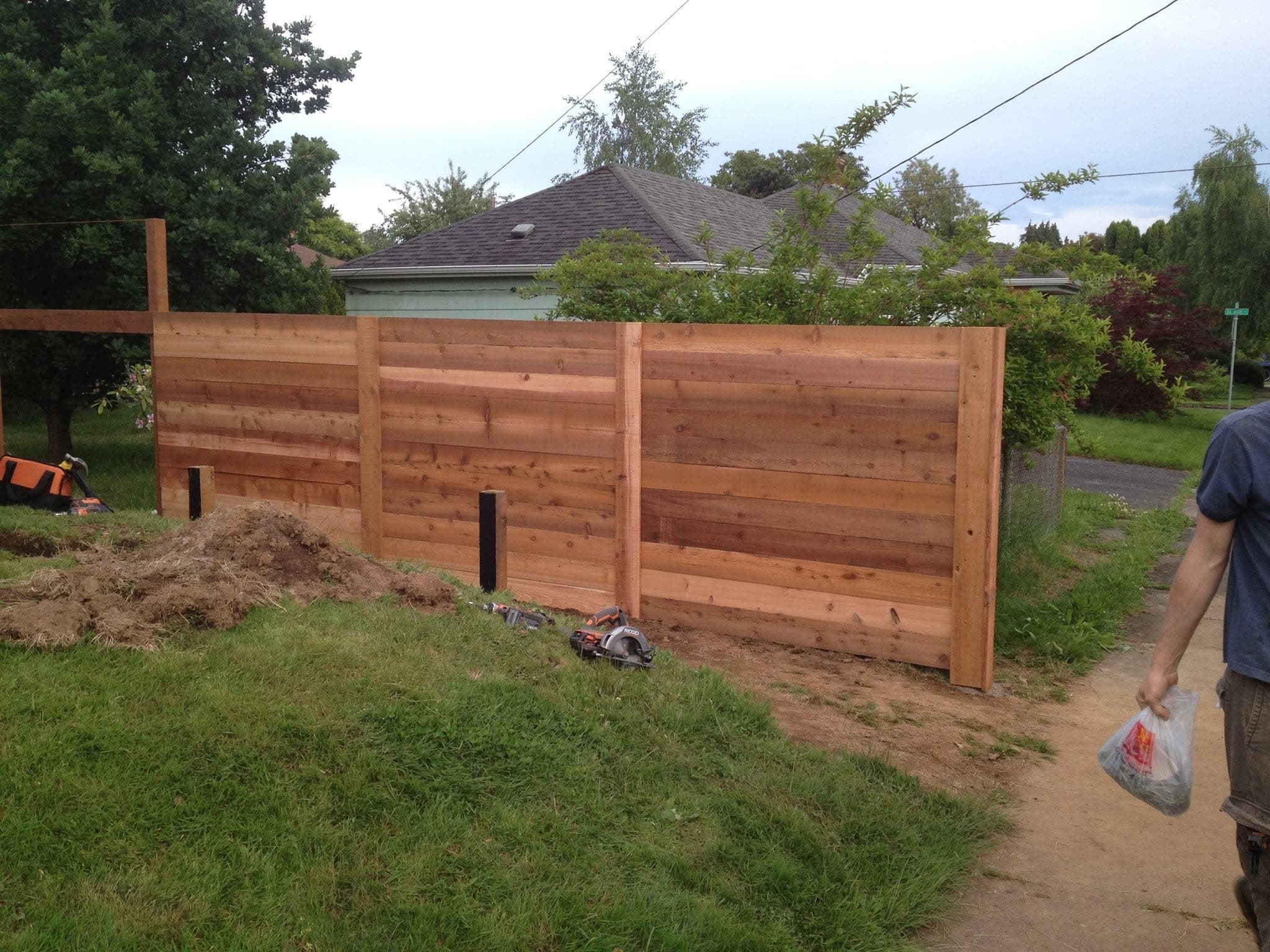 Horizontal-slat cedar fence under construction along a grassy side yard, tools on the ground, worker walking away